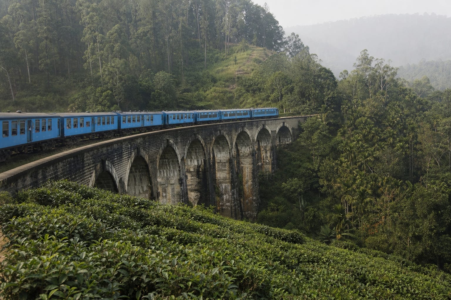 A blue Sri Lankan train crossing a stone viaduct through hill country tea plantations near Ella